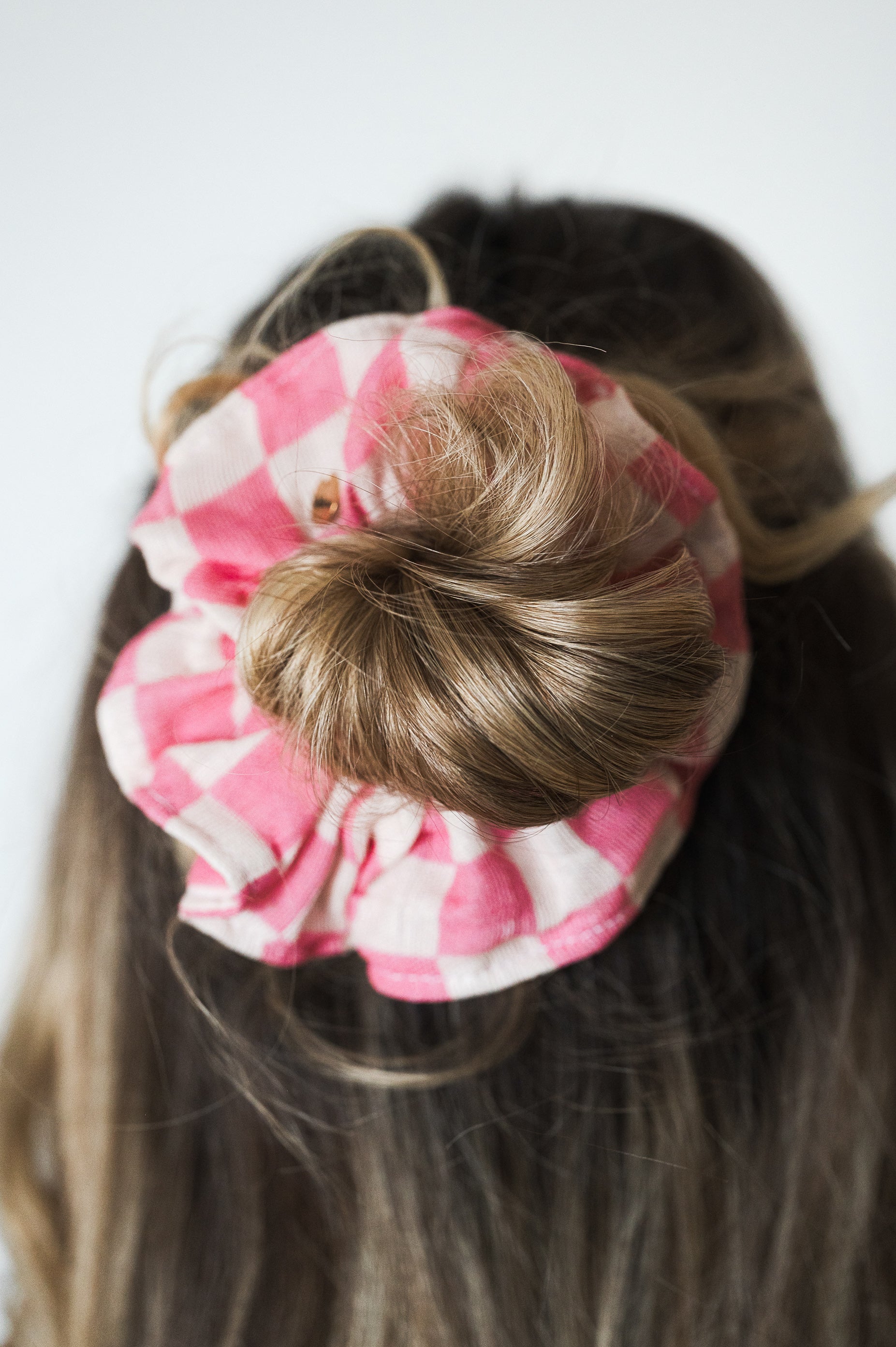 La photo montre un chignon de cheveux avec un chouchou à motif vichy rose et blanc. Les cheveux semblent être de couleur châtain clair. La photo est prise de dos et on ne voit pas le visage de la personne.