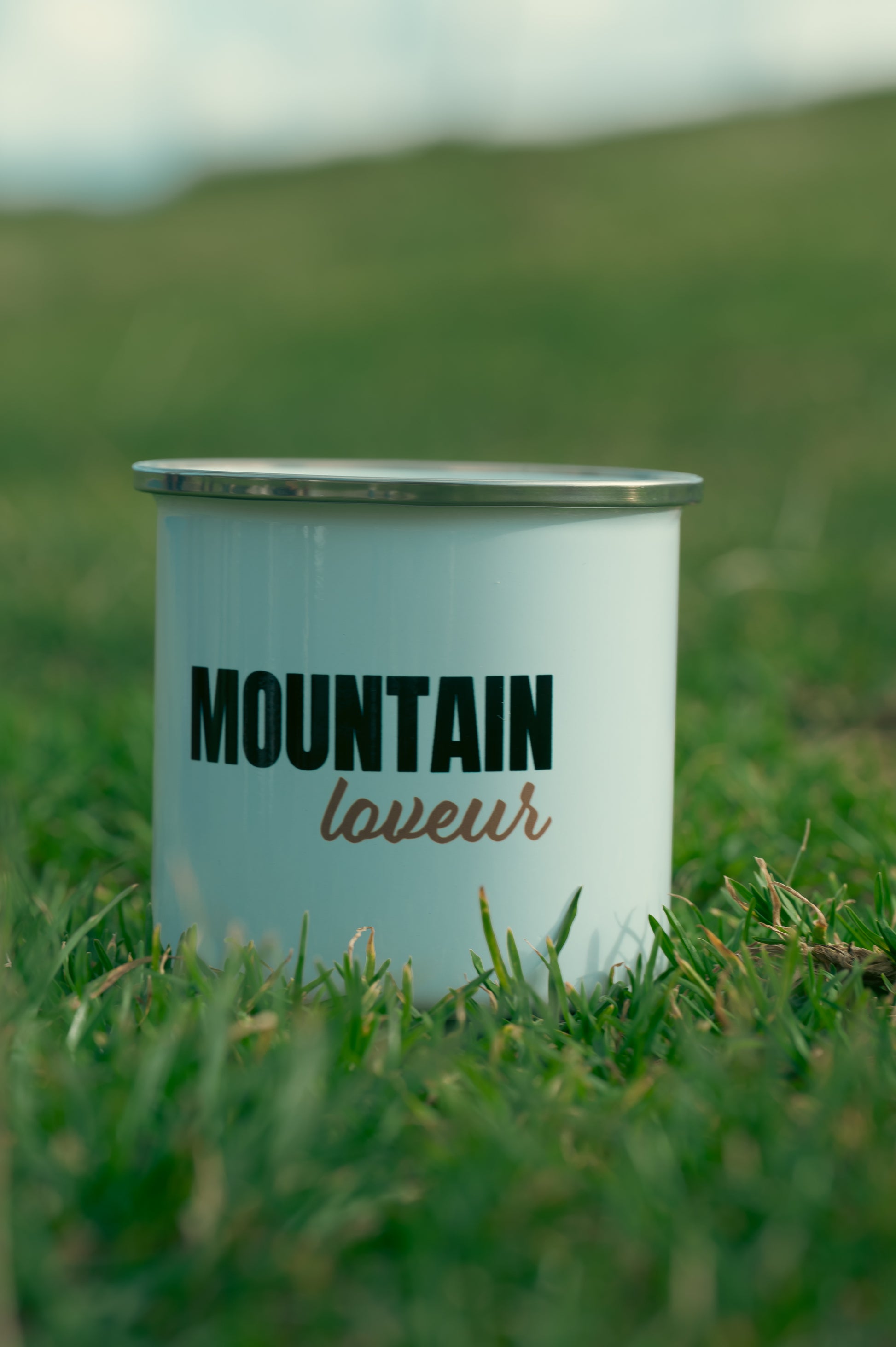 La photo montre un mug posé sur de l'herbe verte. Sur le mug, il y a l'inscription "MOUNTAIN lover" en lettres noires. Le mug semble être de couleur bleu clair avec le bord supérieur en argent ou en métal. En arrière-plan, l'image est floue, mais on peut deviner un paysage verdoyant.