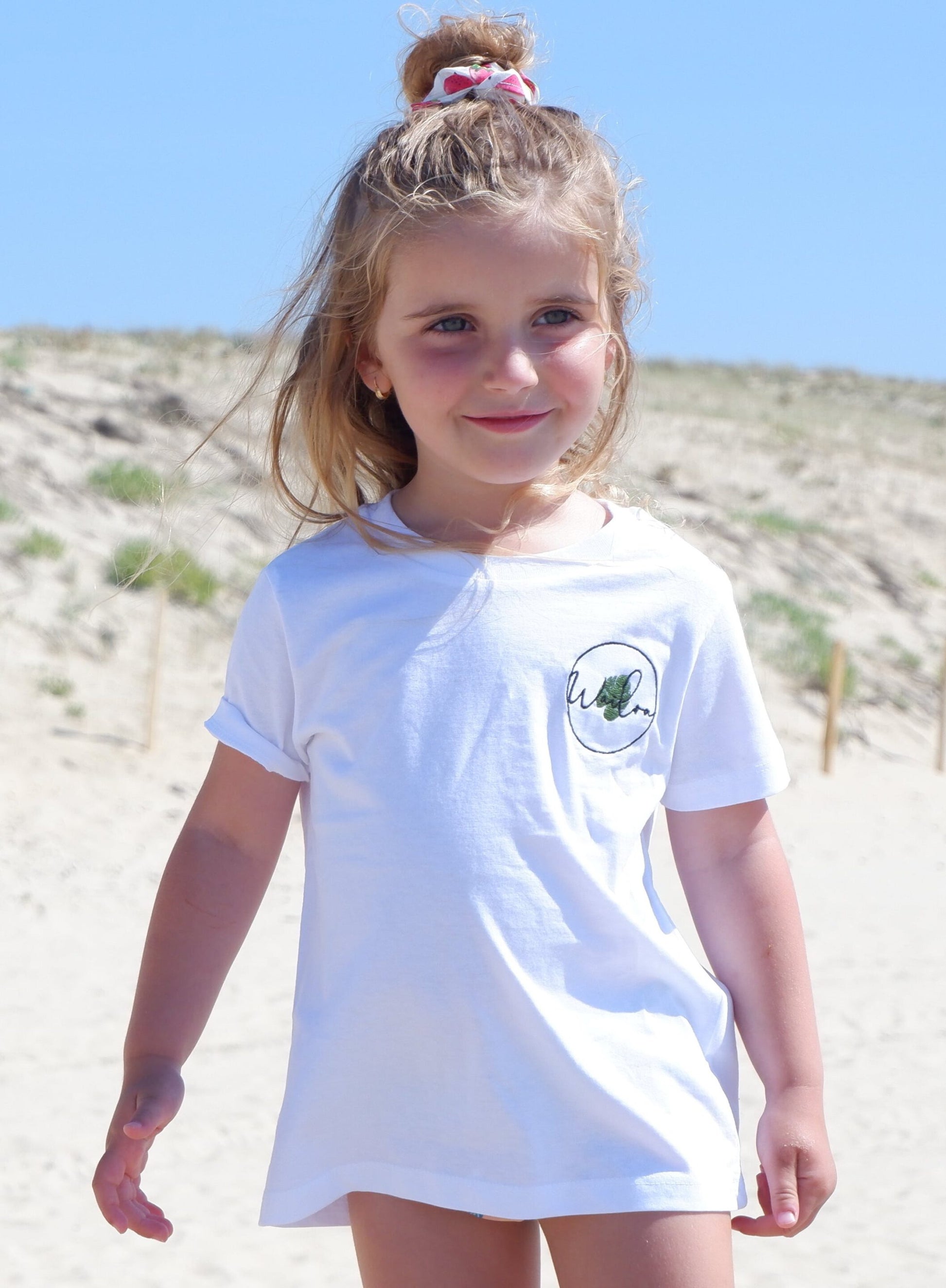 Il s'agit d'une photo d'une petite fille debout sur une plage. Elle porte un t-shirt blanc avec un logo brodé sur le côté gauche de la poitrine. Ses cheveux blonds sont partiellement attachés en haut de sa tête avec un élastique rose. Elle sourit et regarde vers l'appareil photo. Derrière elle, on peut voir un paysage de dunes de sable.