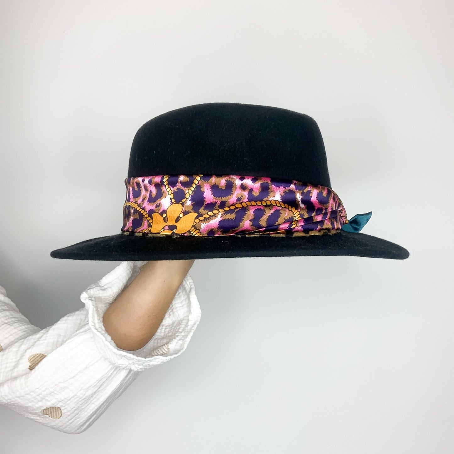 La photo montre un chapeau noir avec un ruban coloré autour de la calotte. Le ruban a un motif animalier avec des taches de couleurs violet, jaune et rose. Le chapeau est posé sur un mannequin ou un support qui a un bras visible vêtu d'une chemise blanche avec des motifs de cœurs marron.