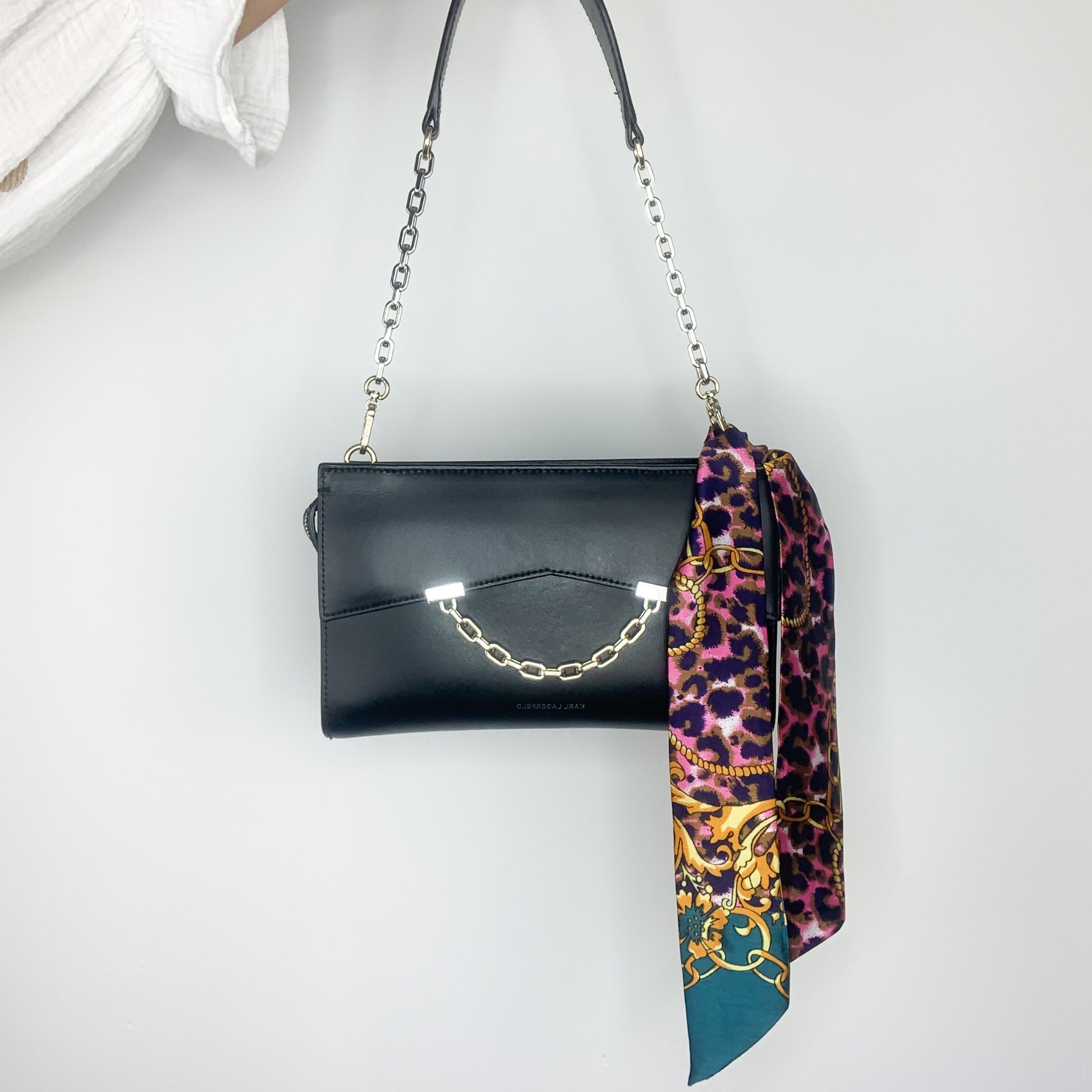 Il s'agit d'une photo d'un sac à main noir avec une chaîne en métal comme bandoulière. Le sac est posé sur une surface blanche et il est ouvert, montrant une doublure intérieure avec un motif léopard dans des tons de violet et jaune.